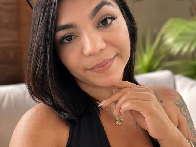 ValeryPierce with medium brown skin, black shoulder-length hair, and a small tattoo on her right arm, smiling, wearing a black halter top, holding a butterfly necklace.