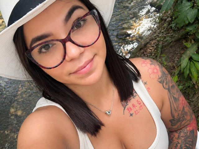 ValeryPierce with dark hair, glasses, white hat, and white tank top showing her tits, has tattoos on her right arm and chest, smiling, standing near a rocky stream.