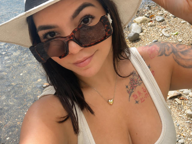 ValeryPierce with dark hair, tortoiseshell sunglasses, white hat, white tank top, chest tattoos, heart necklace, smiling, showing her tits, tattoos on right shoulder, standing by water.