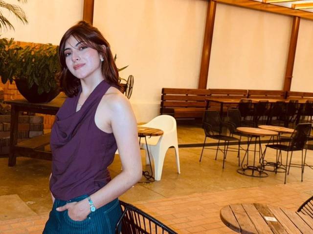 SpencerLe has shoulder-length brown hair, a purple sleeveless top, and blue pinstriped pants, standing with hands in pockets, looking at camera, in a warmly lit outdoor café.
