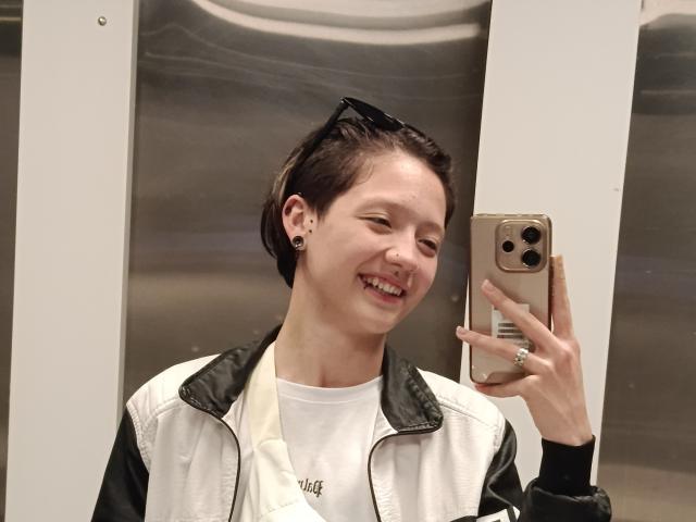 LissyLuxe with short brown hair, black ear gauges, smiling, wearing a white and black jacket, holding a gold iPhone, standing in a metallic room.