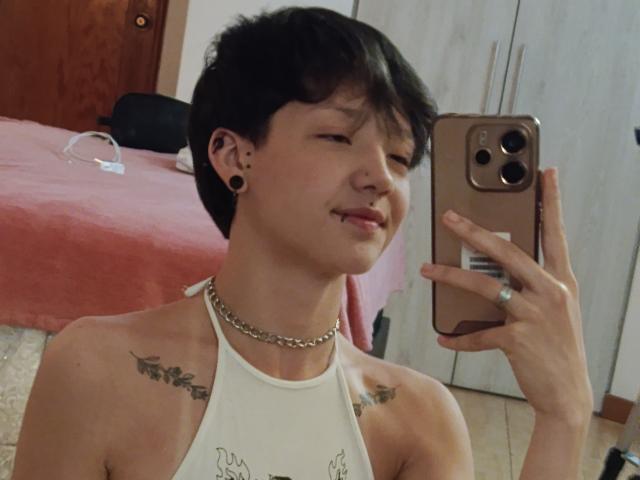LissyLuxe met kort zwart haar, oorbel piercings, een ketting, een wit tanktopje dat kleine tatoeages laat zien, die een selfie maakt in een spiegel, een gouden iPhone vasthoudend, met een lichte grijns..