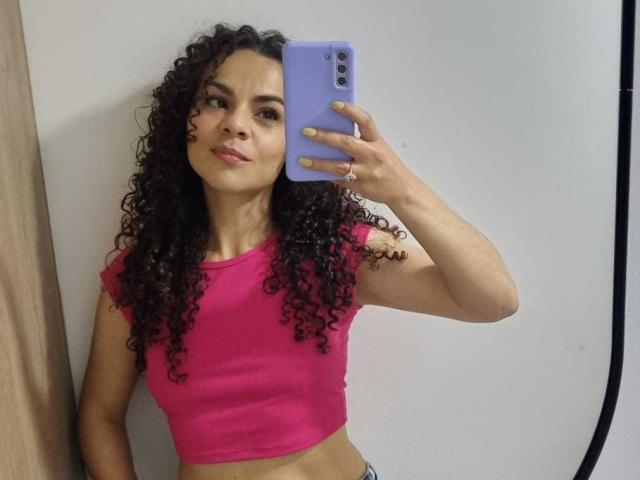 MissSunnyShine with curly dark hair, pink crop top, and light skin, holding a purple phone, taking a mirror selfie, showing her small tits and slightly visible ass.