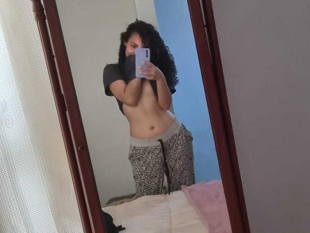 Mirrored selfie of MissSunnyShine with curly black hair, wearing a black shirt lifted to expose her bare tits, and leopard print pants, holding a phone in her right hand.