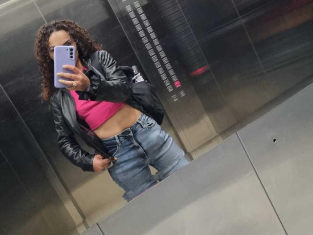 MissSunnyShine in an elevator, wearing a black leather jacket, pink top, and blue jeans, with curly brown hair, showing her belly button with a phone in hand.