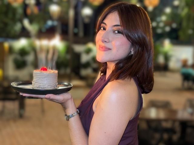 SpencerLe with shoulder-length brown hair, wearing a purple sleeveless top, holding a small cake with a cherry on a black tray, smiling slightly, in a dimly-lit room.