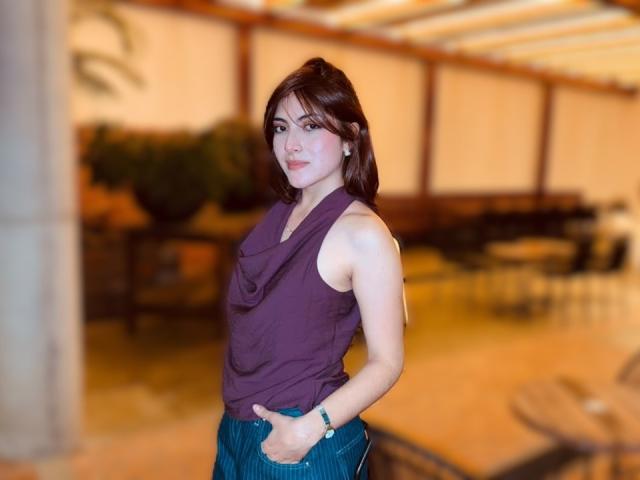 SpencerLe with short brown hair, wearing a purple sleeveless top and blue jeans, hands in pockets, smiling, standing in a blurry indoor setting, looking at camera.