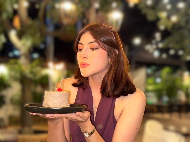SpencerLe with brown hair, pink lips, and purple sleeveless top, blowing out candle on small cake, holding black plate, wearing watch, in dimly lit outdoor setting.