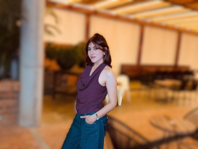SpencerLe with brown hair, wearing a purple sleeveless top and blue pinstriped pants, standing with hands in pockets, looking at camera. Soft focus, indoor setting.