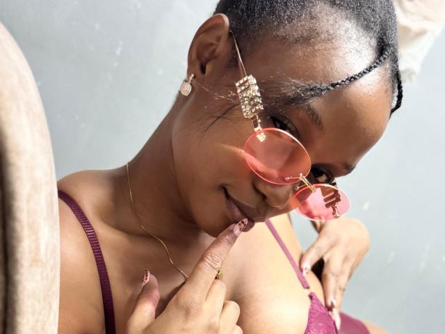 CutyKeshin with dark skin, braided hair, pink sunglasses, gold earrings, and a pink top, touching her finger to her lips, showing off her small, perky tits.