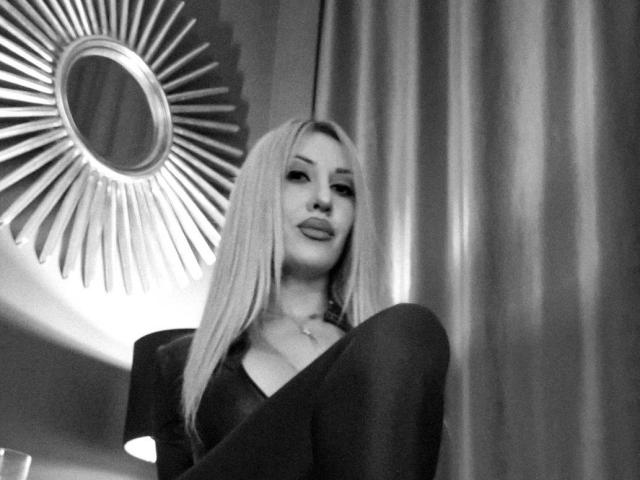 Black-and-white photo of VickiSpices with long blonde hair, sitting, wearing a dark dress, knees up, subtle smile, sunburst mirror in background, soft light, elegant.