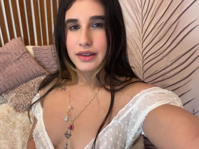 KatieDesirX, avec ses longs cheveux foncés, portant un haut blanc à pois, dévoilant sa poitrine avec un collier, et un petit tatouage au-dessus de sa poitrine, souriant à la caméra..