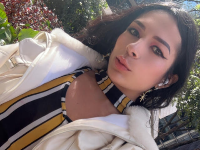 ArielNitzche with pouty lips, wearing a striped top and white jacket, has dark eyebrows, long eyelashes, and star-shaped earrings, looking at the camera in sunlight.