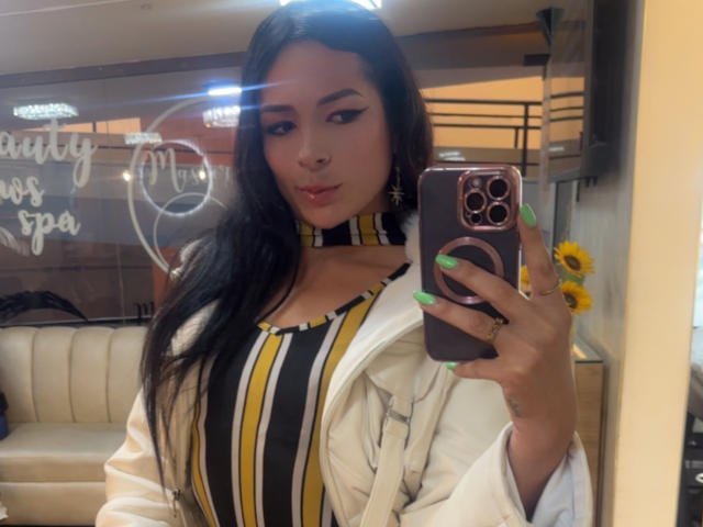 ArielNitzche with long black hair, green nails, and a yellow-striped top, holding a phone, wearing a white jacket, and sunflower earrings, taking a selfie. Boobs visible.