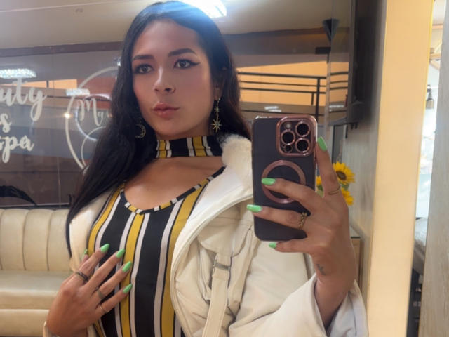 ArielNitzche with green nails, striped yellow-black-brown top, white jacket, holding phone, looking at mirror, brown hair, cute face, earrings, sunflower in background, indoor, casual selfie.