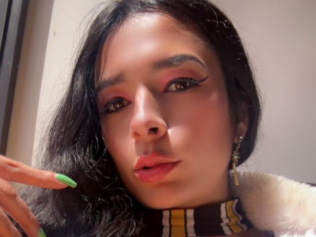 ArielNitzche's face with dark wavy hair, pink eyeshadow, winged eyeliner, green nails, pink lips, fur collar, and a gold earring. She's giving a slight pouty look.