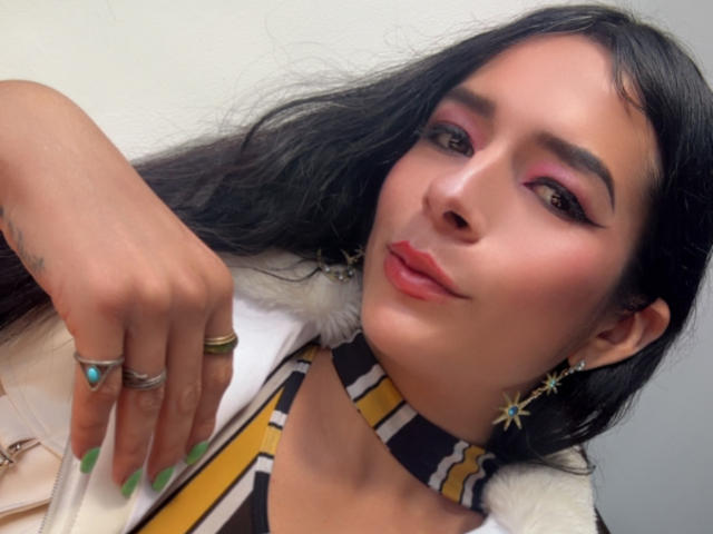 ArielNitzche with pink eyeshadow, green nails, star earrings, and a choker, looking at the camera, wearing a white fur jacket, showing her ringed hand.