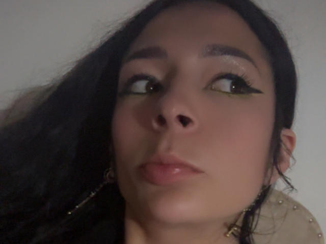 ArielNitzche's face with dark eyes looking up, wearing green eyeliner, black hair, slightly parted lips, and small gold earrings, in a blurry selfie. Her ass is not visible.
