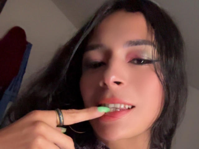 ArielNitzche with dark hair, green nails, and a ring, biting her finger with her pink lipstick and eye makeup. Her finger is near her smiling, slightly open mouth.