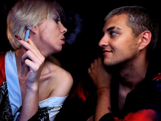 BlondIce, con cabello rubio corto y uñas rojas, sostiene un cigarrillo, con la mirada fija en un hombre de pelo gris que lleva una camisa roja, quien le devuelve la mirada con una ligera sonrisa..