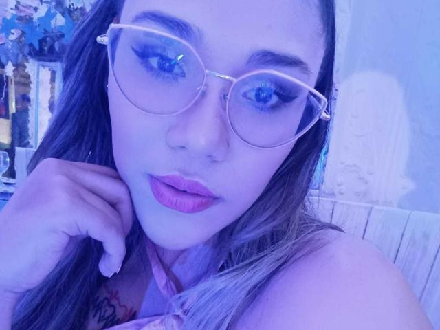 KennyRousse with blue-tinted face, wearing glasses, pink lipstick, and pink top, hand on chin, tattoo on shoulder, long brown hair, and cute, sexy look.