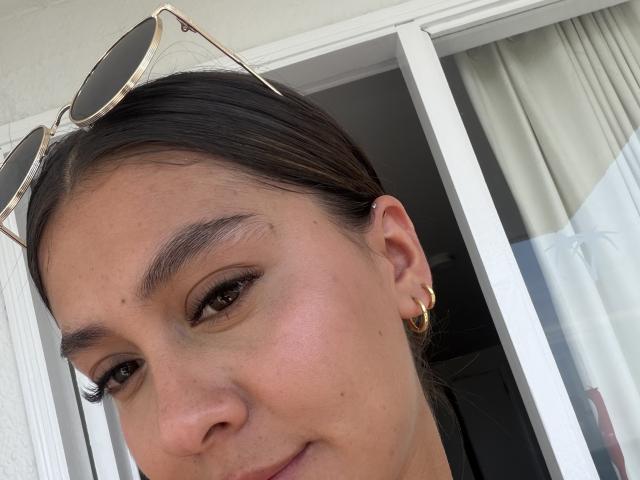 ThaliaAres has brown eyes, a slight smile, and wears gold hoop earrings with sunglasses on her head. Her skin is light with a natural glow.