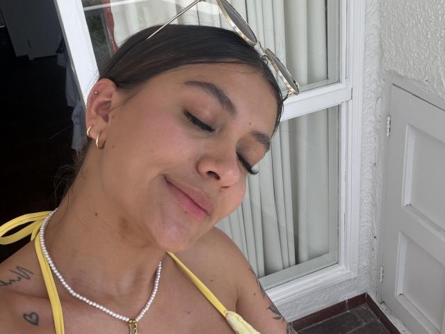ThaliaAres a la peau mate, les yeux fermés et un léger sourire. Elle porte un haut jaune, un collier de perles et des boucles d'oreilles en or, avec des lunettes de soleil sur la tête. On peut voir un tatouage sur son épaule..