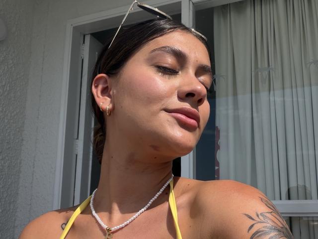 ThaliaAres has tan skin, brown hair, small tattoos, and a yellow halter top. She's wearing sunglasses on her head and a pearl necklace with gold earrings. Sunlight on her face.