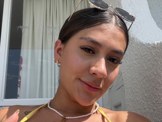 ThaliaAres with tan skin, brown eyes, and a small nose, wearing sunglasses on head, yellow halter top, pearl necklace, and earrings, smiling softly, showing subtle tattoos on neck.