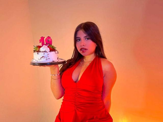 Celeste Morel, com cabelo castanho, vestindo um vestido vermelho, segurando um bolo branco com rosas cor-de-rosa e velas, tem seios grandes e um rosto bonito, em um quarto com iluminação quente..