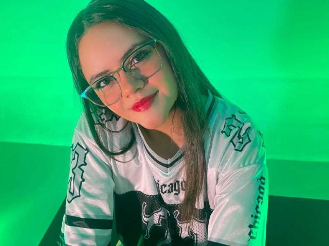 Emily Colsson met een bril, lang bruin haar, rode lippenstift, een wit sportshirt met zwarte en grijze designs, groen licht, voorovergebogen, met een lichte glimlach..