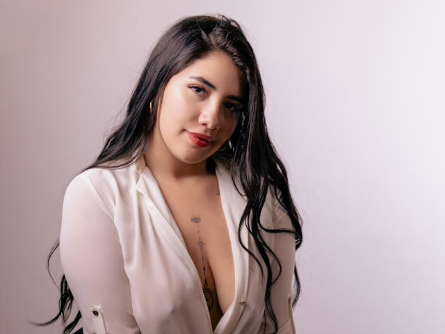 EmaFiore with long black hair, white open shirt, and a small tattoo above her tits, smiling softly. She has a nose ring and red lipstick.