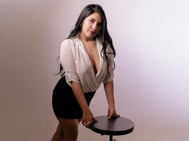 EmaFiore with long black hair, white deep V-neck blouse showing her big boobs, black skirt, leaning on a round table, hands on top, smiling, white background.