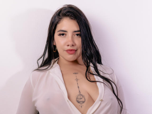 EmaFiore with long black hair, wearing a sheer white button-down, showing large boobs and a chest tattoo, has a slight smile and is against a plain white background.