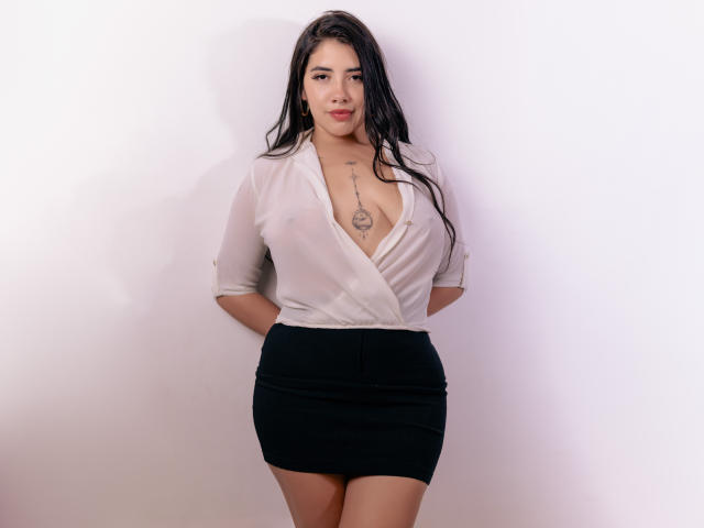 EmaFiore has large tits, a deep V-neck white shirt, black skirt, long black hair, tattoo on chest, hands behind back, standing, white wall background, looking at camera.