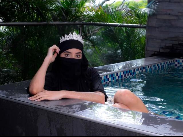 MariamNur in a black mask and crown, black shirt, half-submerged in a blue-tiled hot tub, hand on head, looking at camera, greenery in background, water reflecting light.