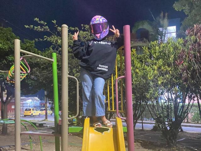 Julia Bennett, con un casco viola, una felpa nera e jeans blu, in piedi su uno scivolo di plastica giallo, con le braccia alzate, in un parco giochi di notte..