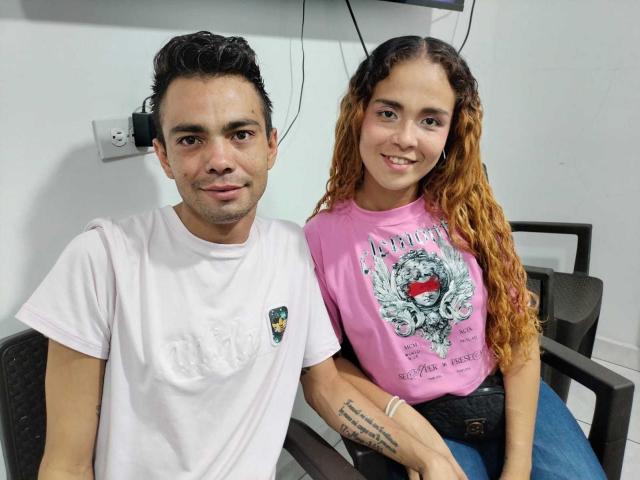 YusAndKleider, con cabello castaño rizado, una camisa rosa y un tatuaje, está sentada al lado de YusAndKleider, con cabello negro, una camiseta blanca y una ligera sonrisa, en una habitación con sillas grises..