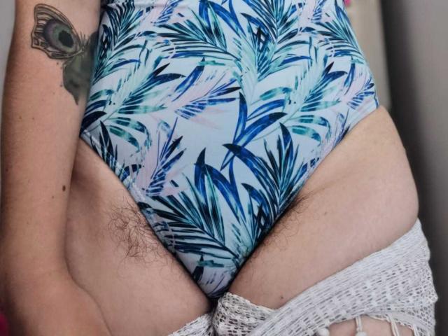 HairyValeri's butt in tight, blue-patterned panties with visible pubic hair, white lace stockings partially down, tattoo of a peacock eye on left upper arm.