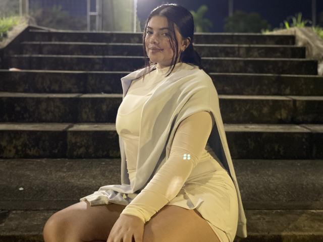 LaraJames, met een groot achterwerk en grote borsten, zittend op betonnen trappen 's nachts, gekleed in een doorschijnende witte jurk, met lange mouwen en een grijze sjaal, kijkend naar de camera..