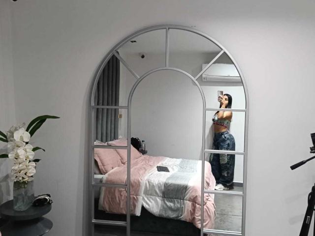 ZammySanz, avec des seins en soutien-gorge, debout devant un miroir, reflétant ses fesses en jean, dans une chambre avec du linge de lit rose et des fleurs blanches..