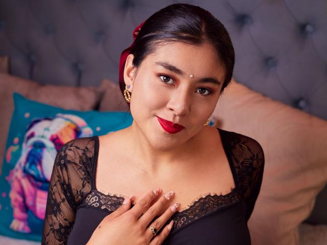 HannaGoez with dark hair in a red hairpiece, wearing a black lace top, red lipstick, and a nose dot, smiling at the camera, hand on chest, tits visible.