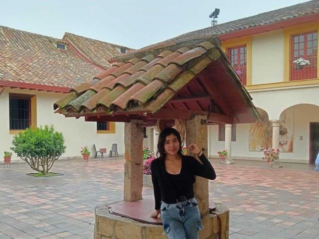 HannaGoez with medium tits, black shirt, and blue jeans, leaning on a tiled roof structure, holding a finger to her mouth, standing in a courtyard with a house.