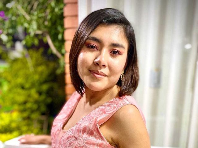 HannaGoez has short brown hair, pink eyeshadow, and a pink, patterned sleeveless top, smiling slightly with a small nose and gold earrings, looking cute and confident.