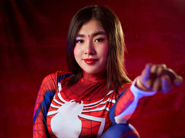 HannaGoez with red spiderweb superhero outfit, pointing forward, red lipstick, brown hair, white spider symbol, blue and red suit, subtle makeup, confident expression, red background.