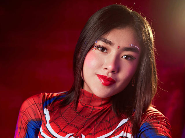 HannaGoez in a Spider-Man costume, red lips, straight black hair, sparkly forehead ornament, wearing red, blue, and white Spider-Man suit, soft smile, slightly flushed cheeks.