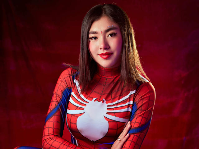 HannaGoez in a red Spider-Man cosplay with white web pattern, crossed arms, and red lipstick, has medium-length brown hair, and a cute, confident expression.