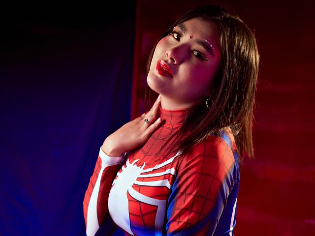 HannaGoez in a Spider-Man cosplay, red and white suit, looking at camera with hand on neck, red lipstick, brown hair, dark eyes, ring on finger, soft light.
