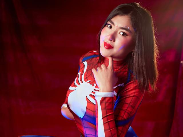 HannaGoez in a Spider-Man cosplay, red and blue suit with white spider, hand on chest, red lipstick, brown hair, cute, looking at camera, background blurred.