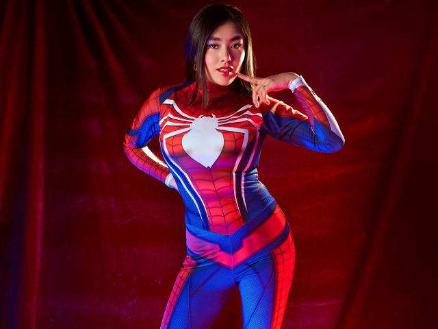 HannaGoez in a Spider-Man costume, red and blue with a white spider, standing with finger to lips, looking sexy, medium tits, tight suit, red background, photo.